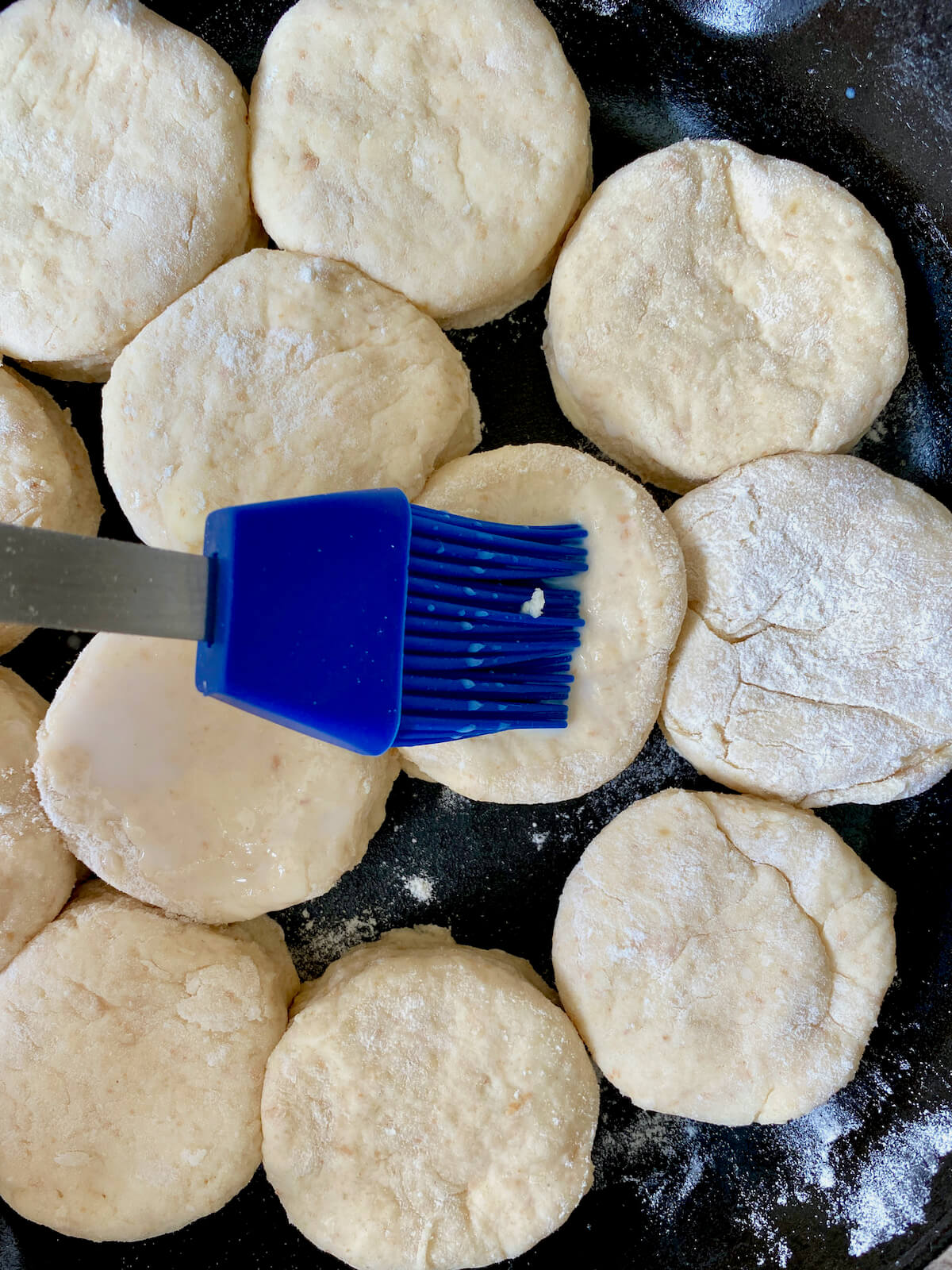 A blue silicone brush brushing buttermilk onto the raw biscuits in the cast iron skillet.