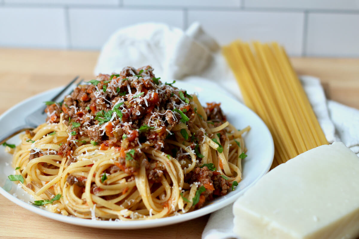 A plate of linguine with bolognese sauce with a fork resting on the side of the plate. The plate is next to a white cloth napkin, dried linguine, and a piece of parmesan.