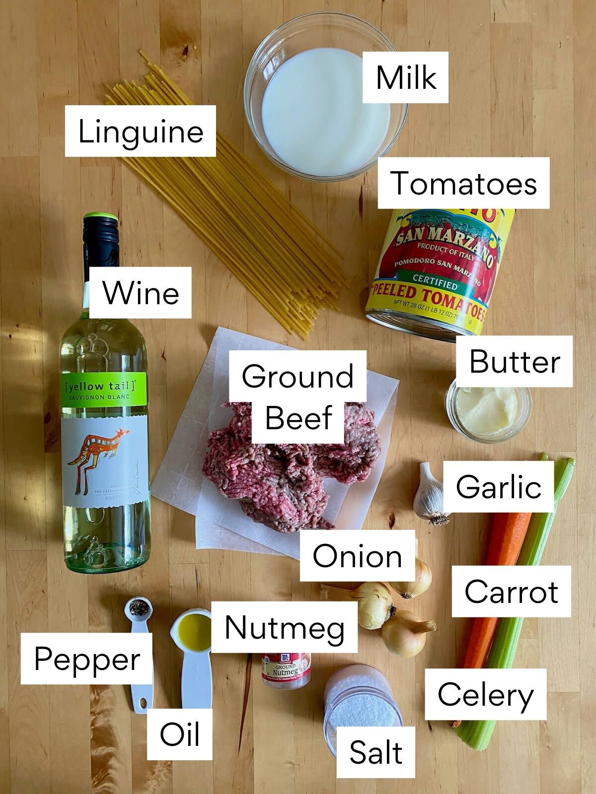 The ingredients to make linguine bolognese on a butcher block countertop. Each ingredient is labeled with text. They include linguine, milk, tomatoes, wine, ground beef, butter, garlic, onion, carrot, celery, nutmeg, pepper, oil, and salt.