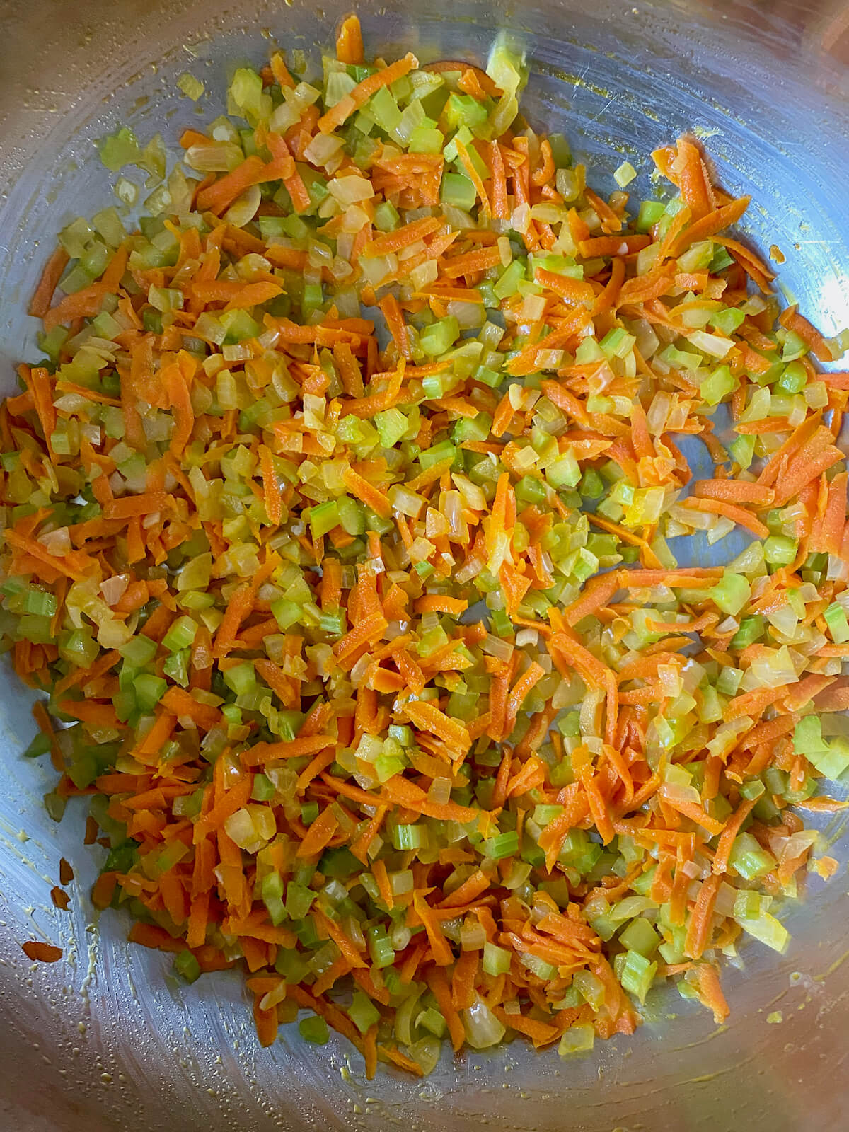 Grated carrots, diced celery, and diced onion sautéing in butter and olive oil in a stainless steel pot.