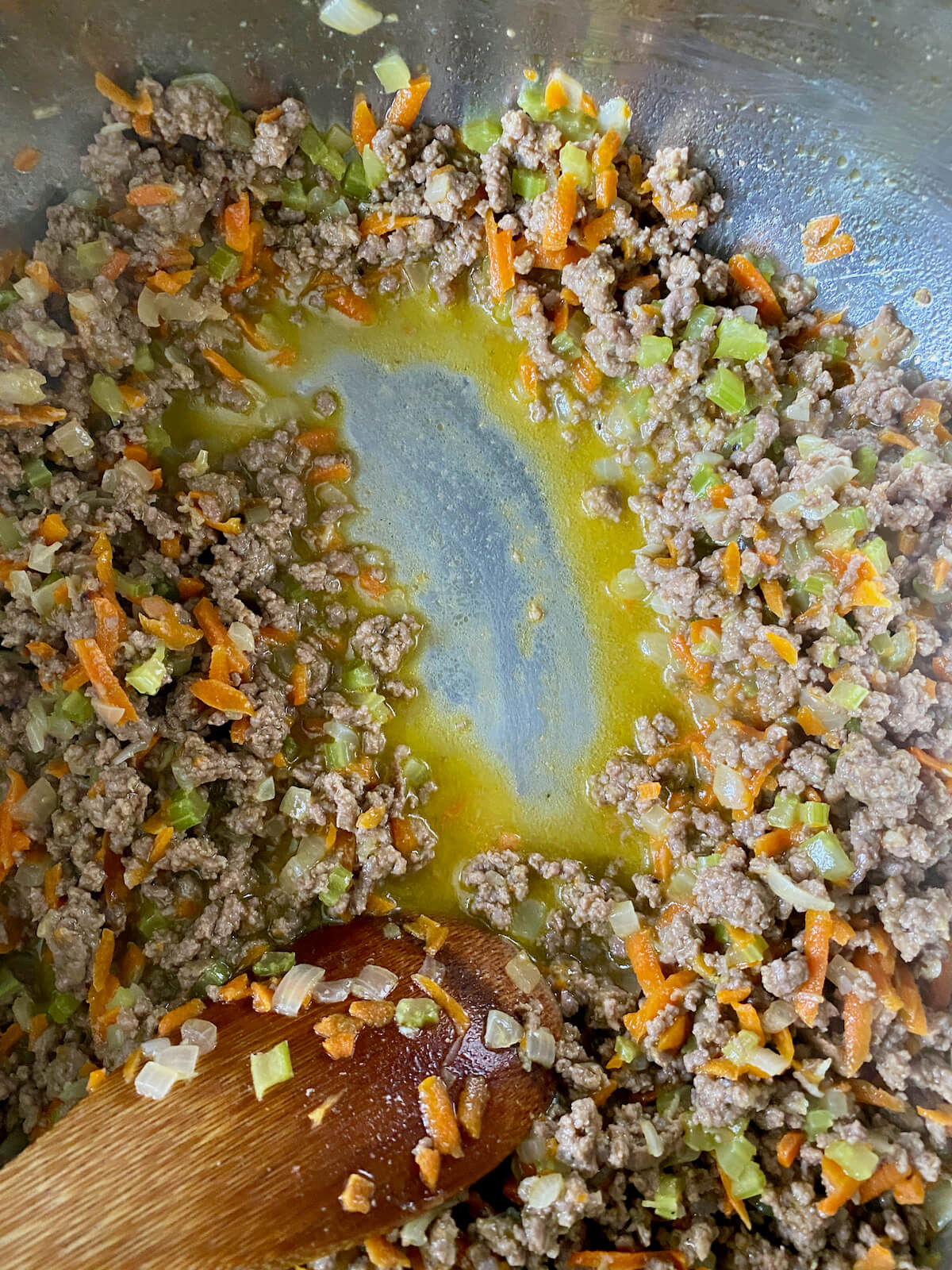 Ground beef and vegetables after simmering in milk. There is a wooden spoon pushing some of the ground beef mixture aside to show that the milk has been reduced in the pan.
