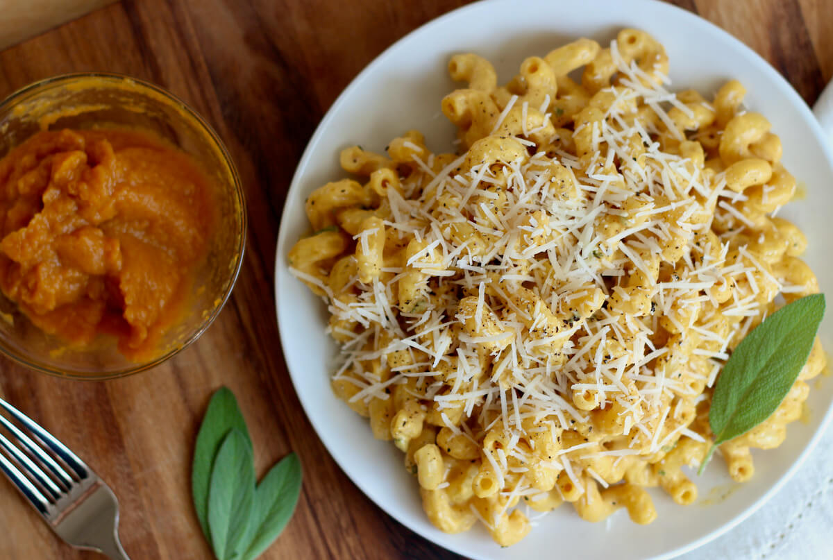 A plate of pumpkin pasta sauce on cavatappi pasta next to a small bowl of pumpkin puree, sage leaves, and a fork.