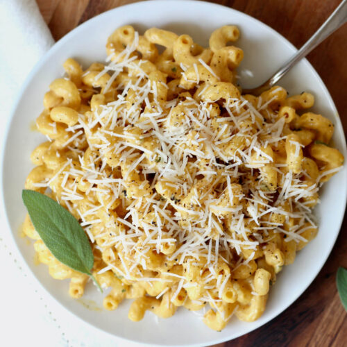 A small white plate of creamy pumpkin pasta sauce on cavatappi pasta. The pasta is garnished with grated parmesan cheese, black pepper, and one whole sage leaf.