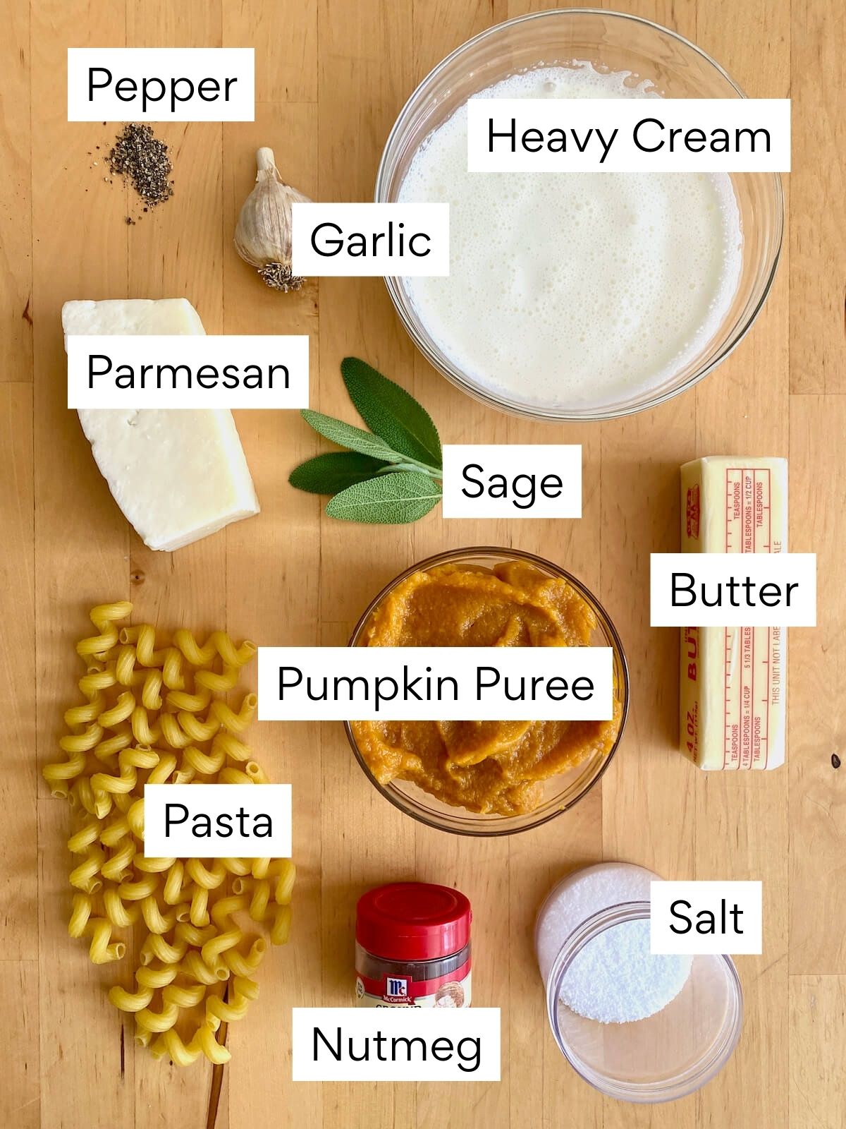 The ingredients to make pumpkin pasta sauce. Each ingredient is labeled with text. They include pepper, garlic, heavy cream, parmesan, sage, butter, pumpkin puree, pasta, nutmeg, and salt.