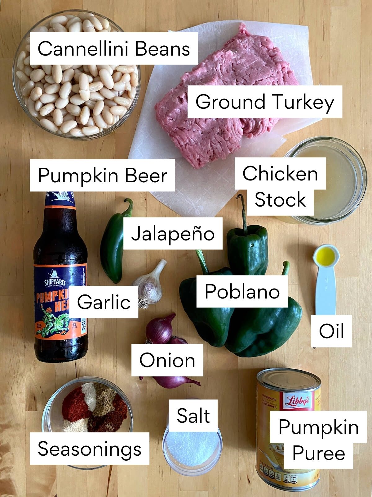 The ingredients to make pumpkin white bean chili with ground turkey. Each ingredient is labeled with text. They include cannellini beans, ground turkey, pumpkin beer, chicken stock, jalapeño, garlic, poblano, onion, oil, seasonings, salt, pumpkin puree.