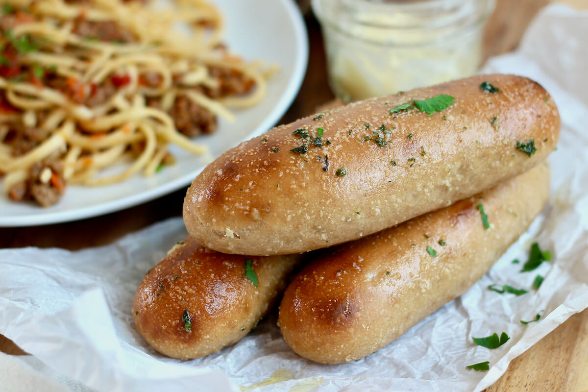 Garlic sourdough breadsticks garnished with herbs. In the background is a plate of pasta with sauce and a jar of butter.