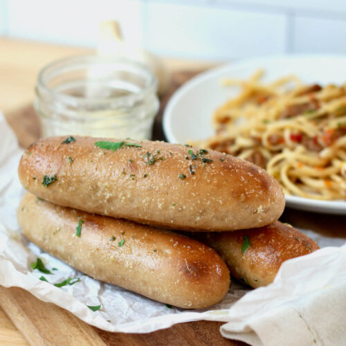 Three sourdough bread sticks stacked on top of each other. In the background is a plate of pasta and a jar of butter.