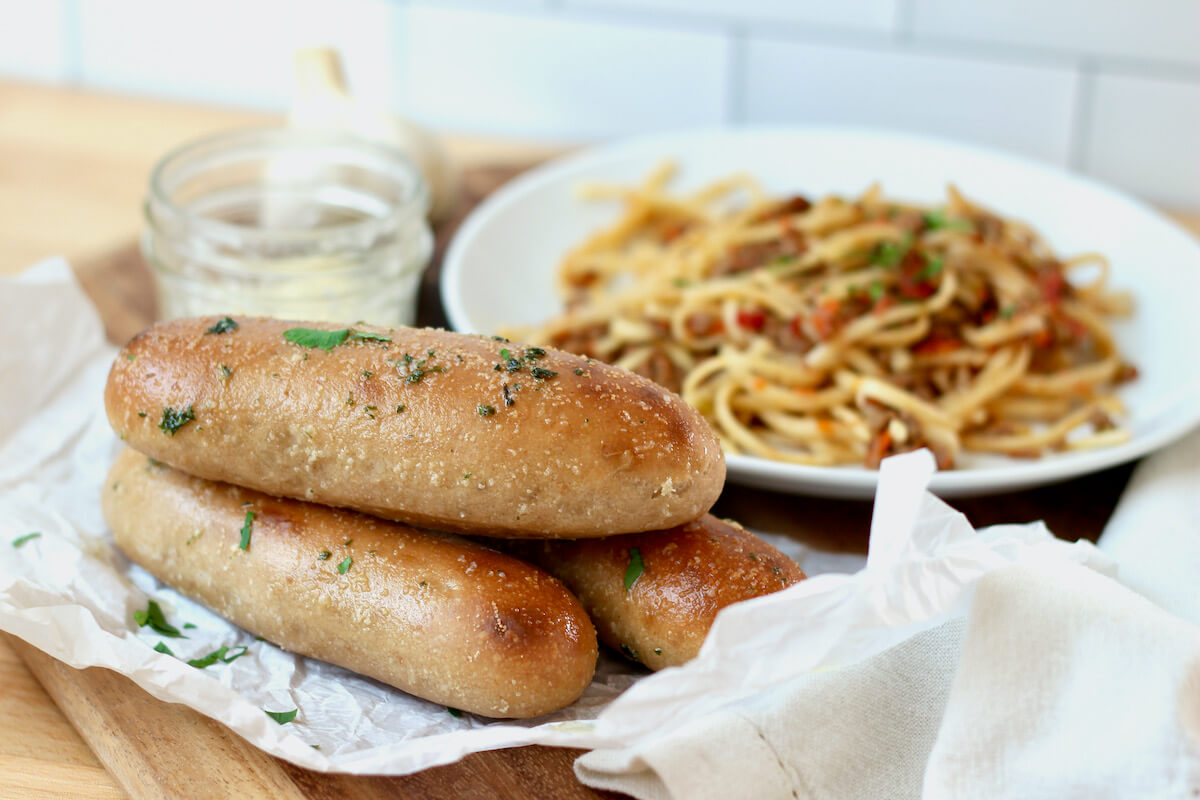 Three sourdough bread sticks stacked on top of each other. In the background is a plate of pasta and a jar of butter.