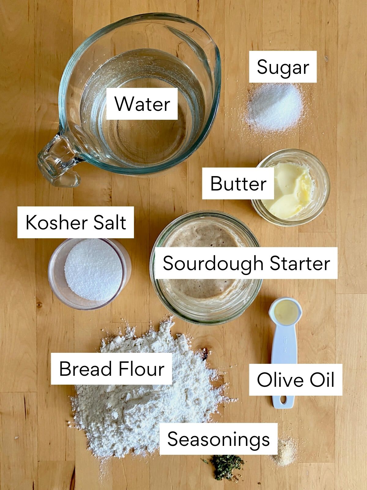The ingredients to make sourdough breadsticks. Each ingredient is labeled with text. They include water, sugar, kosher salt, butter, sourdough starter, bread flour, olive oil, and seasonings.