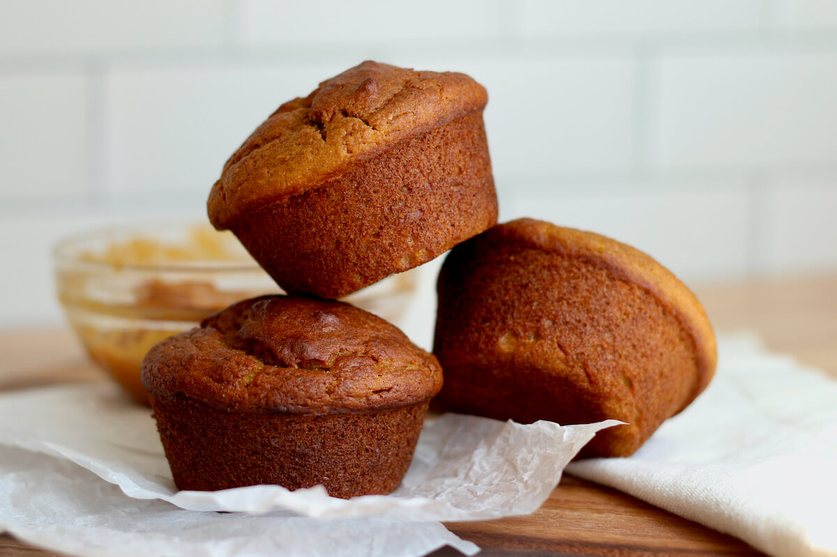 Three sourdough pumpkin muffins stacked in a pyramid shape on a wrinkled piece of parchment paper. There is a small bowl of pumpkin puree in the background.