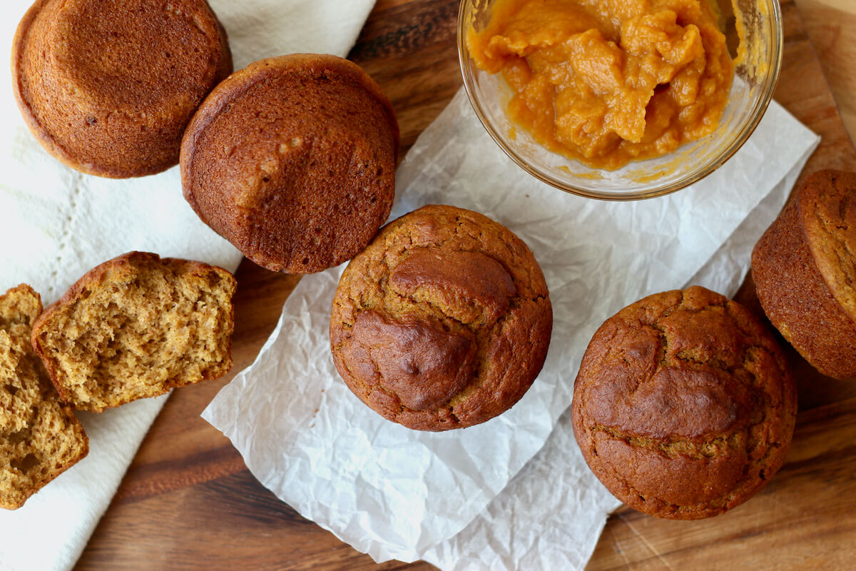 Several sourdough pumpkin muffins laying across a wooden countertop next to a small bowl of pumpkin puree. One of the muffins is split in half down the middle on the left side.