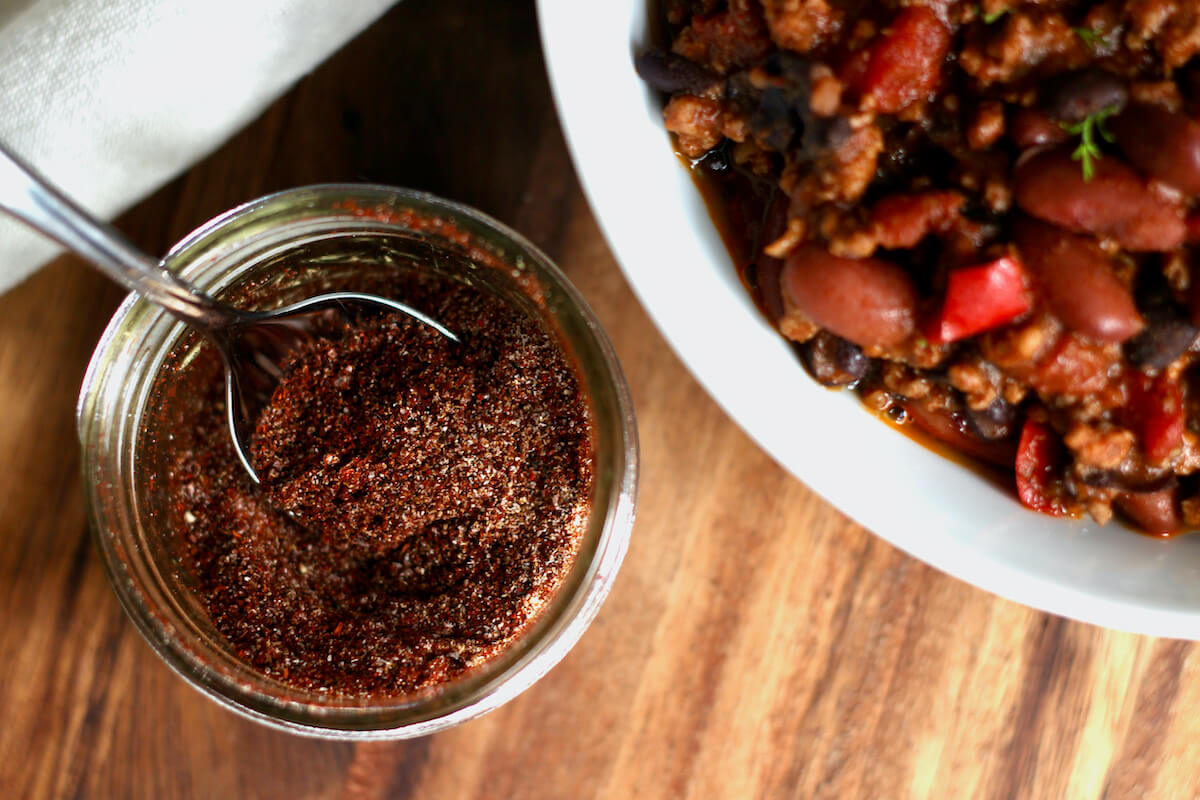 A small glass jar of chili seasoning with a spoon in it next to a bowl of chili.