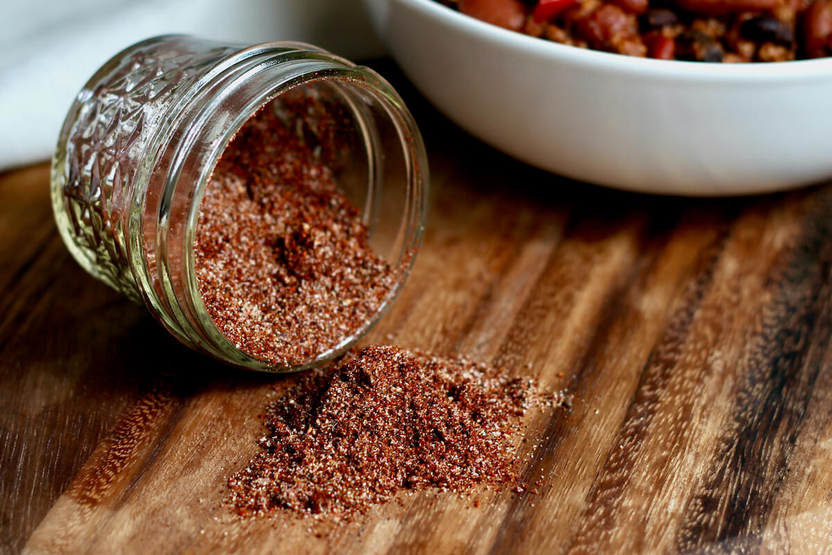 A small glass jar of chili seasoning tipped over with some of the seasoning spilling out. Out of focus in the background is a bowl of chili.