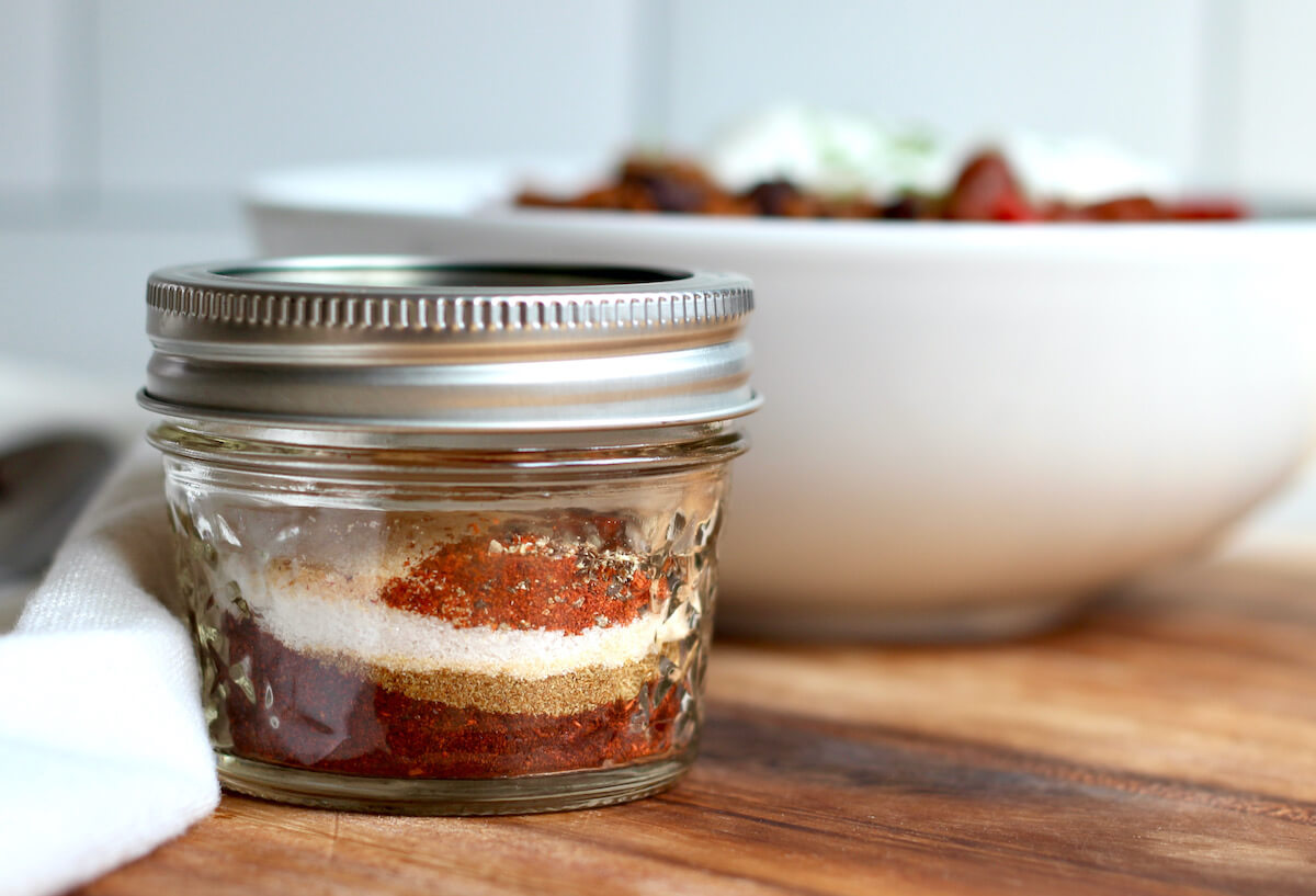 A small glass jar of chili seasoning showing each individual spice in layers before being mixed together. There is a bowl of chili in the background.