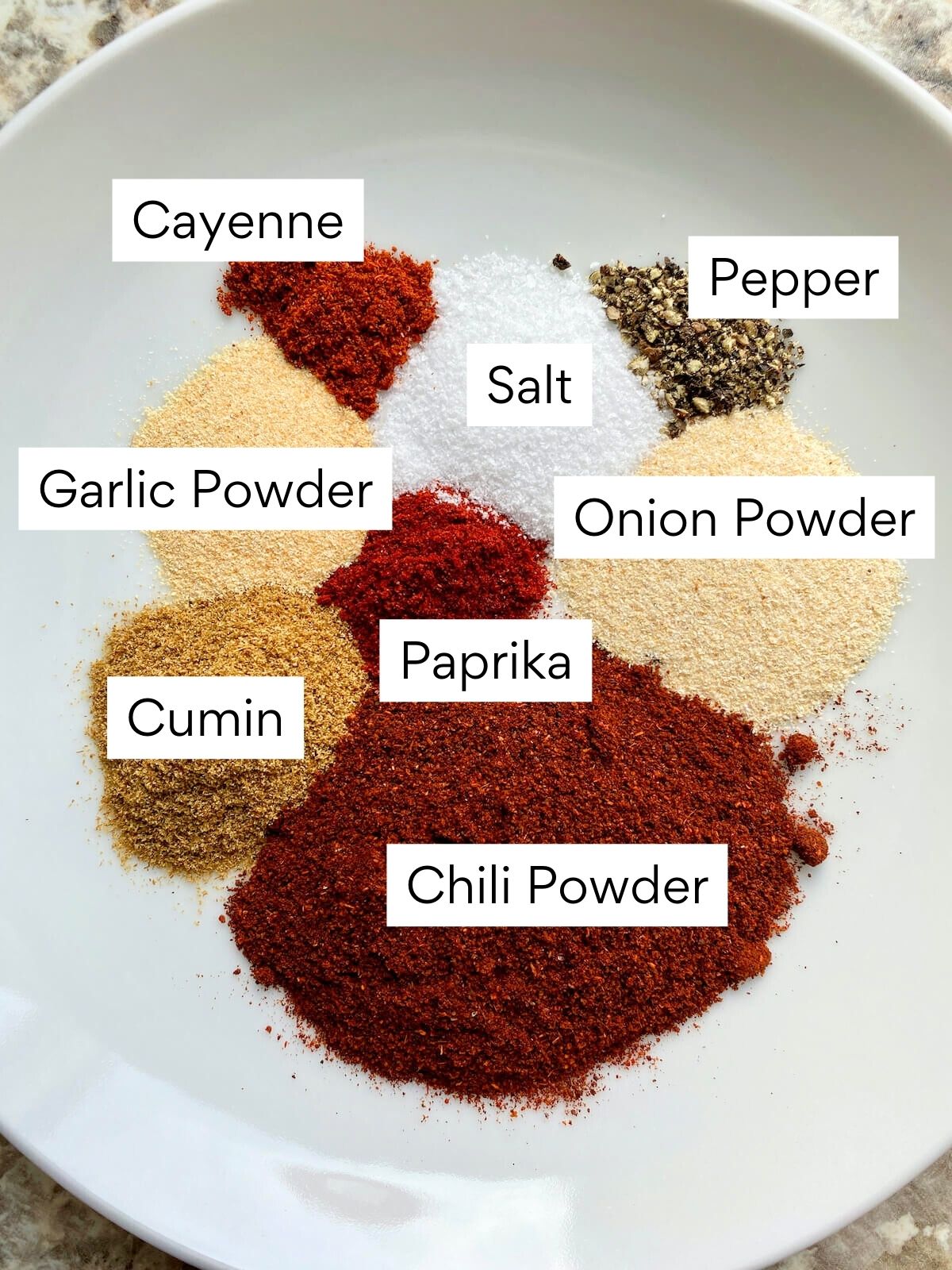 The ingredients to make homemade chili seasoning. Each ingredient is labeled with text. They include chili powder, cumin, garlic powder, paprika, onion powder, cayenne, salt, and pepper.