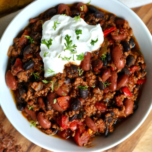 A bowl of Dutch oven chili topped with sour cream and cilantro. The dish is next to a small jar of chili seasoning and a piece of cornbread.