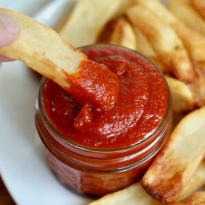 A plate of fries with a small jar of homemade ketchup. There's a hand dipping a fry into the ketchup.