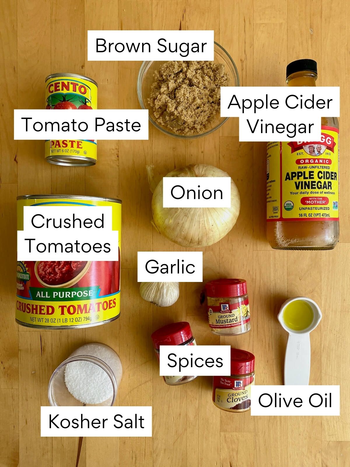The ingredients to make homemade ketchup. Each ingredient is labeled with text. They include tomato paste, crushed tomatoes, brown sugar, apple cider vinegar, onion, garlic, spices, kosher salt, and olive oil.
