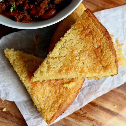 Three slices of old-fashioned cornbread stacked on a piece of parchment paper next to a bowl of chili.