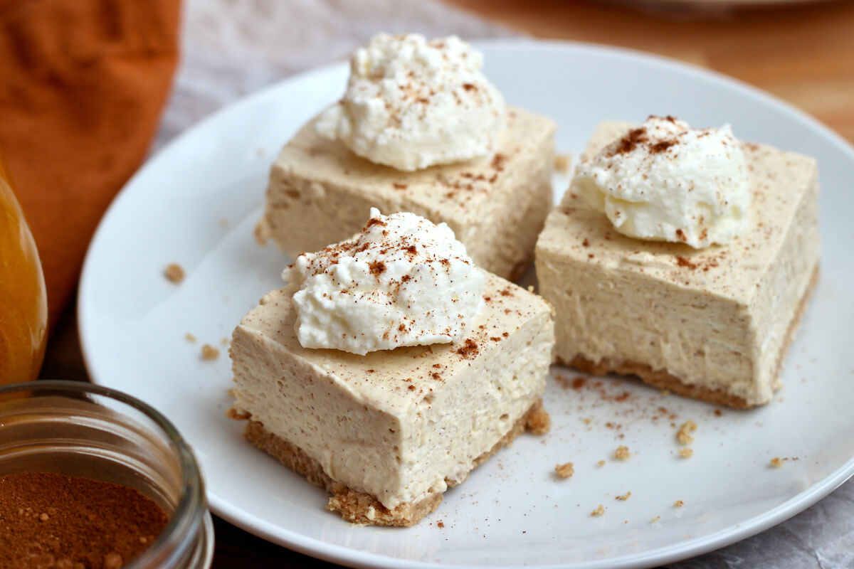 Three no-bake pumpkin cheesecake bars on a small white plate. Each bar is topped with a dollop of whipped cream and sprinkled with pumpkin pie spice.