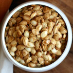 A small white bowl of roasted pumpkin seeds on a wooden countertop next to orange and white cloth napkins.