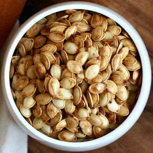 A small white bowl of roasted pumpkin seeds on a wooden countertop next to orange and white cloth napkins.