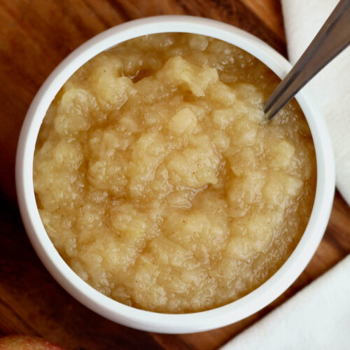 A small white bowl filled with unsweetened applesauce. There is a spoon sticking out of the applesauce to the right. Next to the bowl is a white cloth napkin.