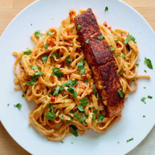 A large white dinner plate with a pile of Cajun alfredo pasta with a piece of blackened salmon on top.