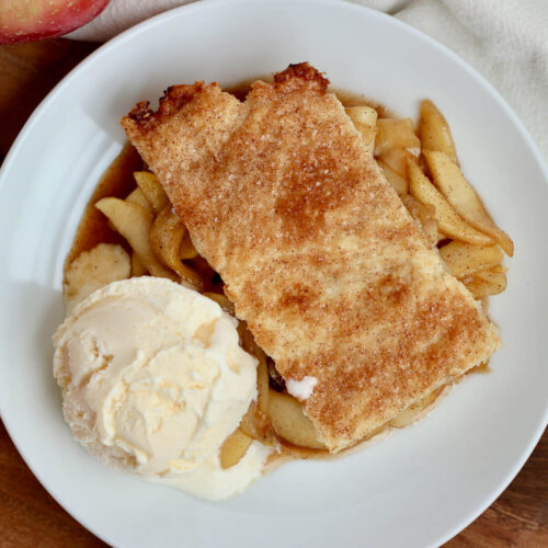 Deconstructed apple pie and a scoop of vanilla ice cream on a small white plate. There is an apple and a white cloth napkin next to the plate.