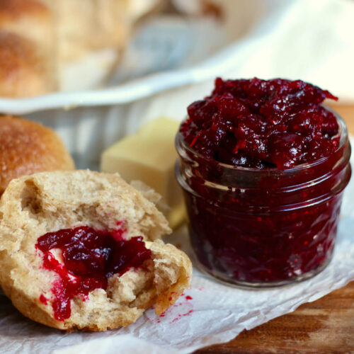 A small jar of cranberry jam next to some dinner rolls and a half stick of butter. One of the rolls has butter and jam on it.