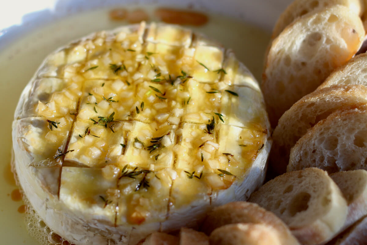 A wheel of baked Brie topped with a garlic honey and herb mixture. There are small slices of bread next to the cheese wheel.
