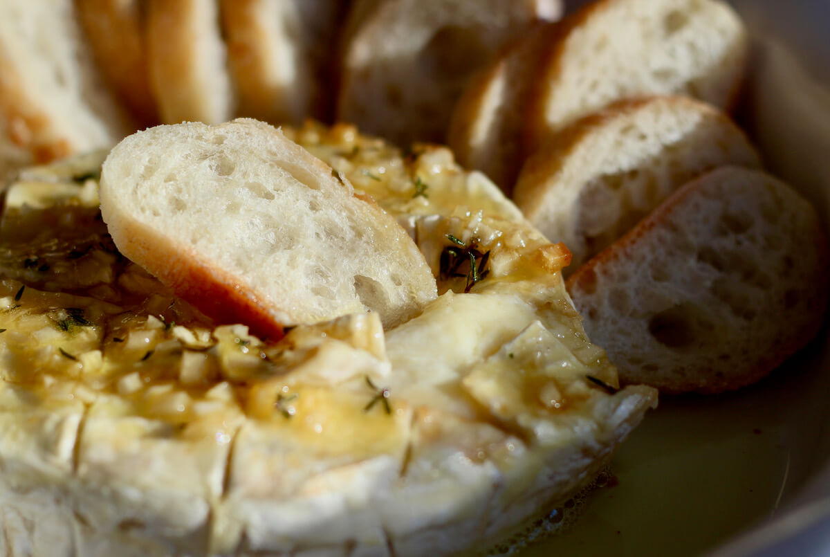 A small slice of bread resting in a wheel of warm baked Brie with garlic, honey, and thyme.
