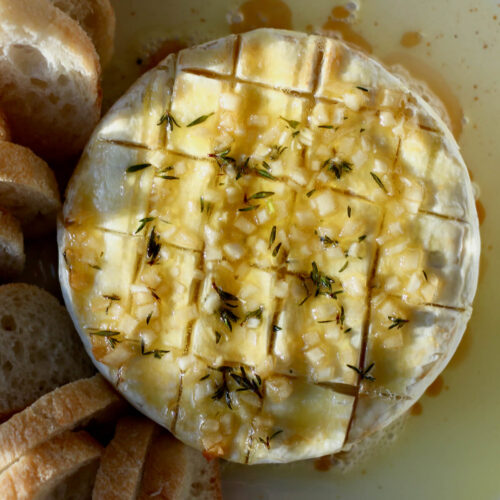 Baked Brie with garlic and herbs in a white pie dish surrounded by olive oil and small slices of bread.