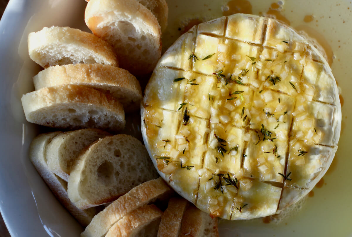 Baked Brie with garlic and herbs in a white pie dish surrounded by olive oil and small slices of bread.