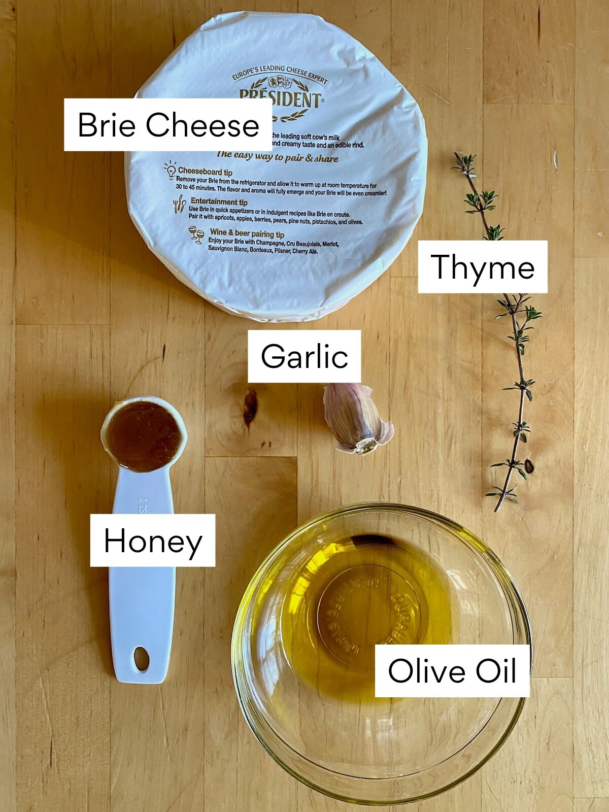The ingredients to make baked brie with garlic. Each ingredient is labeled with text. They include Brie cheese, thyme, garlic, honey, and olive oil.