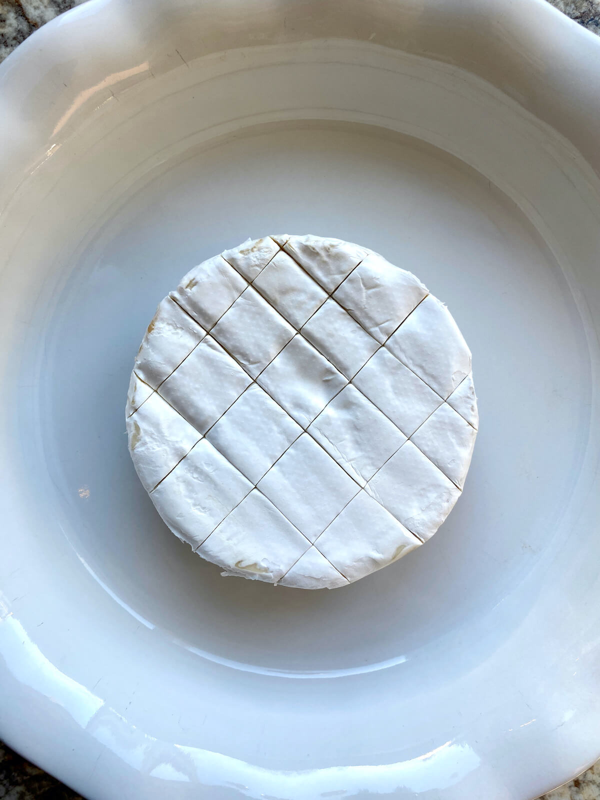 A wheel of Brie cheese with cuts through the top rind in a pie dish.
