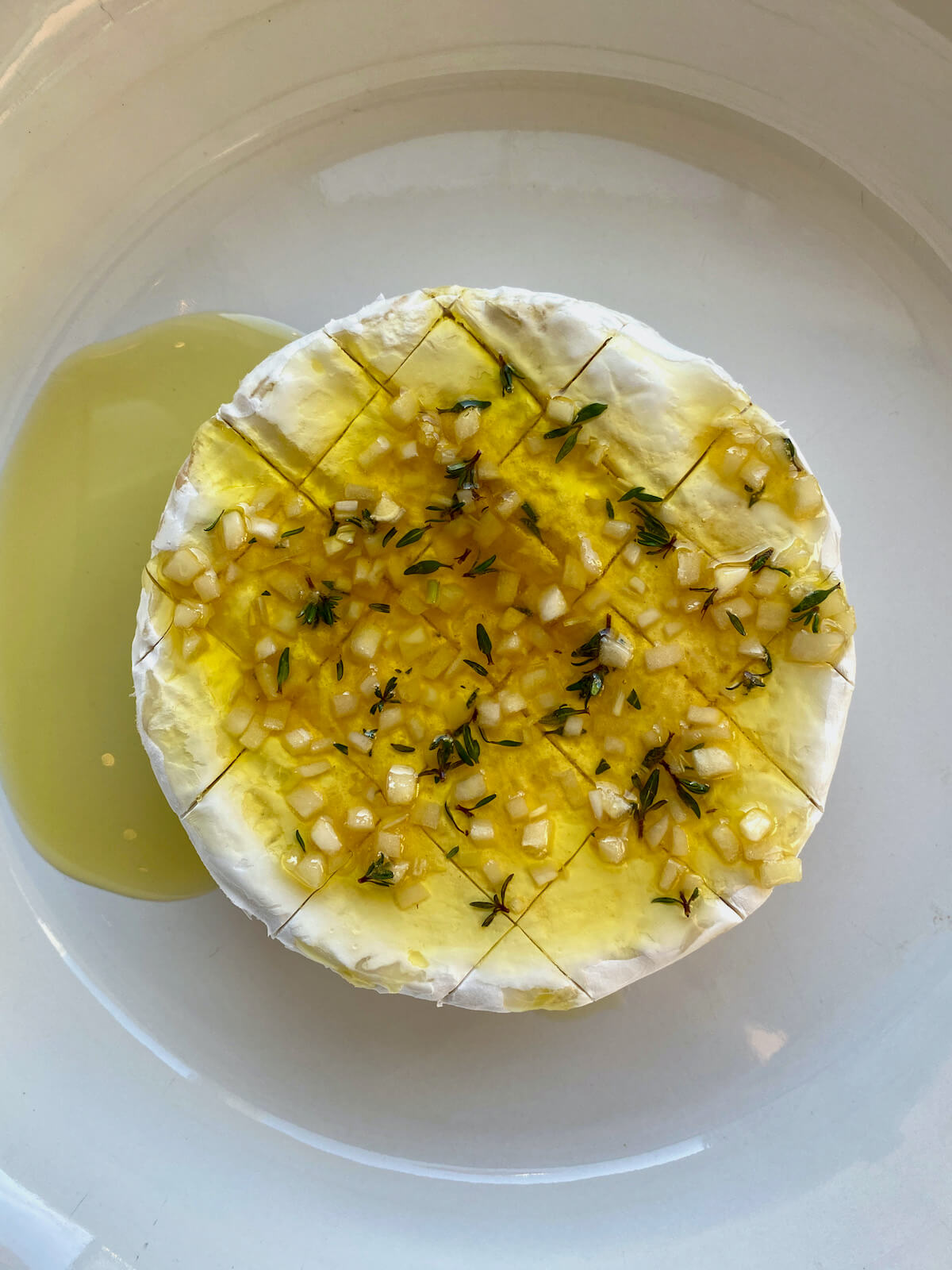 Brie cheese topped with olive oil, honey, garlic, and thyme in a white pie plate.