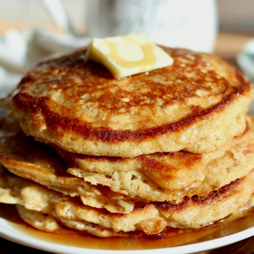 A stack of five sourdough buttermilk pancakes with a pat of butter and maple syrup drizzled over the top.