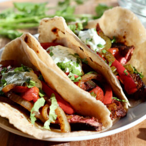 Three beef fajita tacos on a small white plate topped with cilantro garlic sauce.
