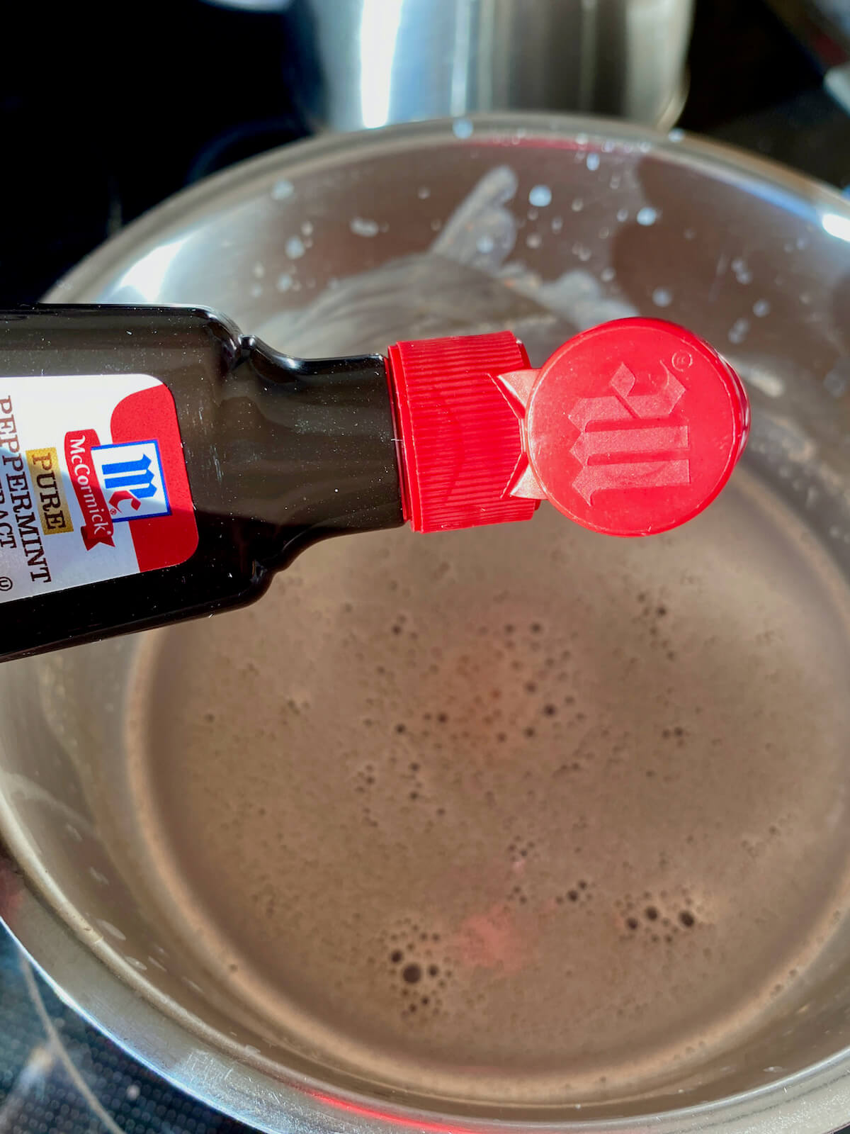 A bottle of peppermint extract being held above a saucepan filled with homemade hot chocolate.