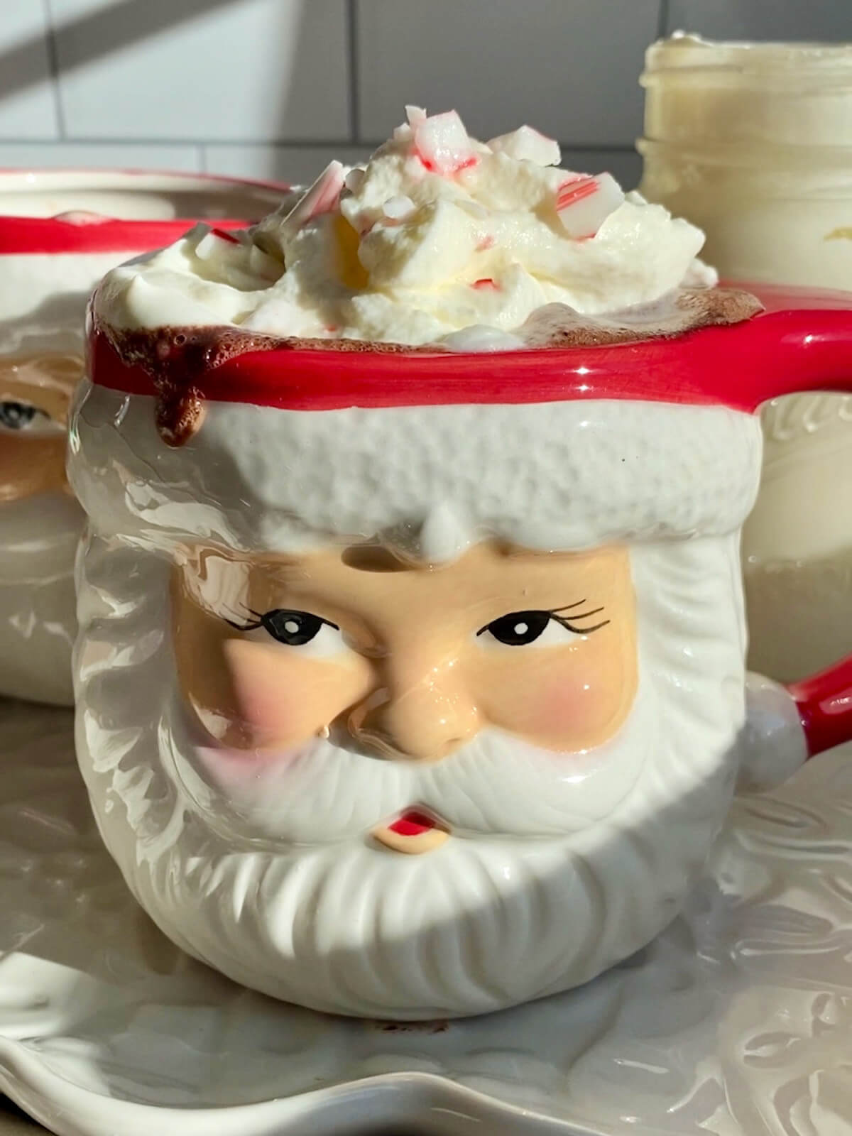 A Santa mug brimming with homemade peppermint hot chocolate. The hot chocolate is topped with homemade whipped cream and crushed candy cane pieces.
