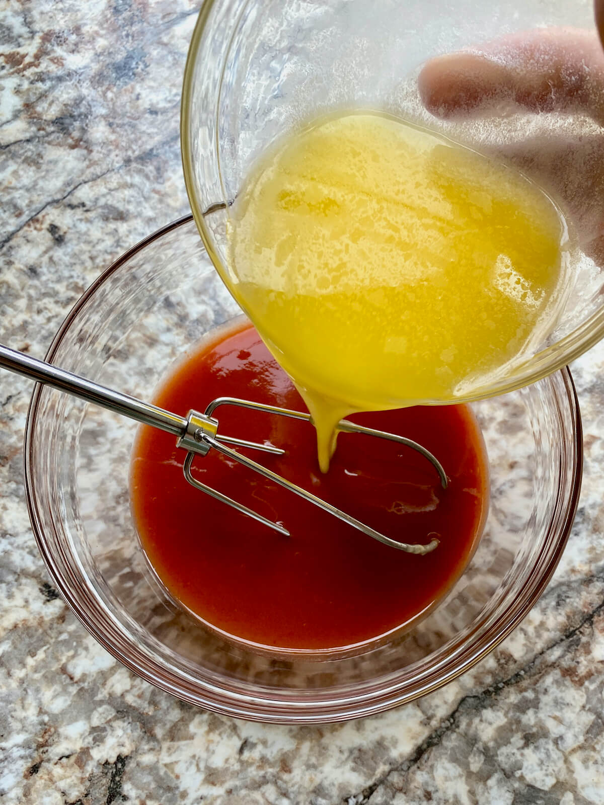 A small bowl of melted butter being poured into a slightly large bowl of cayenne pepper hot sauce. There is a small whisk in the bowl of hot sauce.