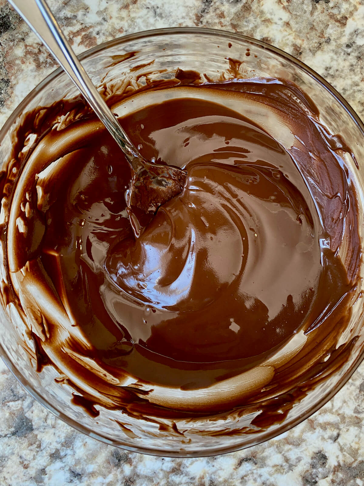 A clear glass bowl filled with melted chocolate with a spoon sticking out to the left.