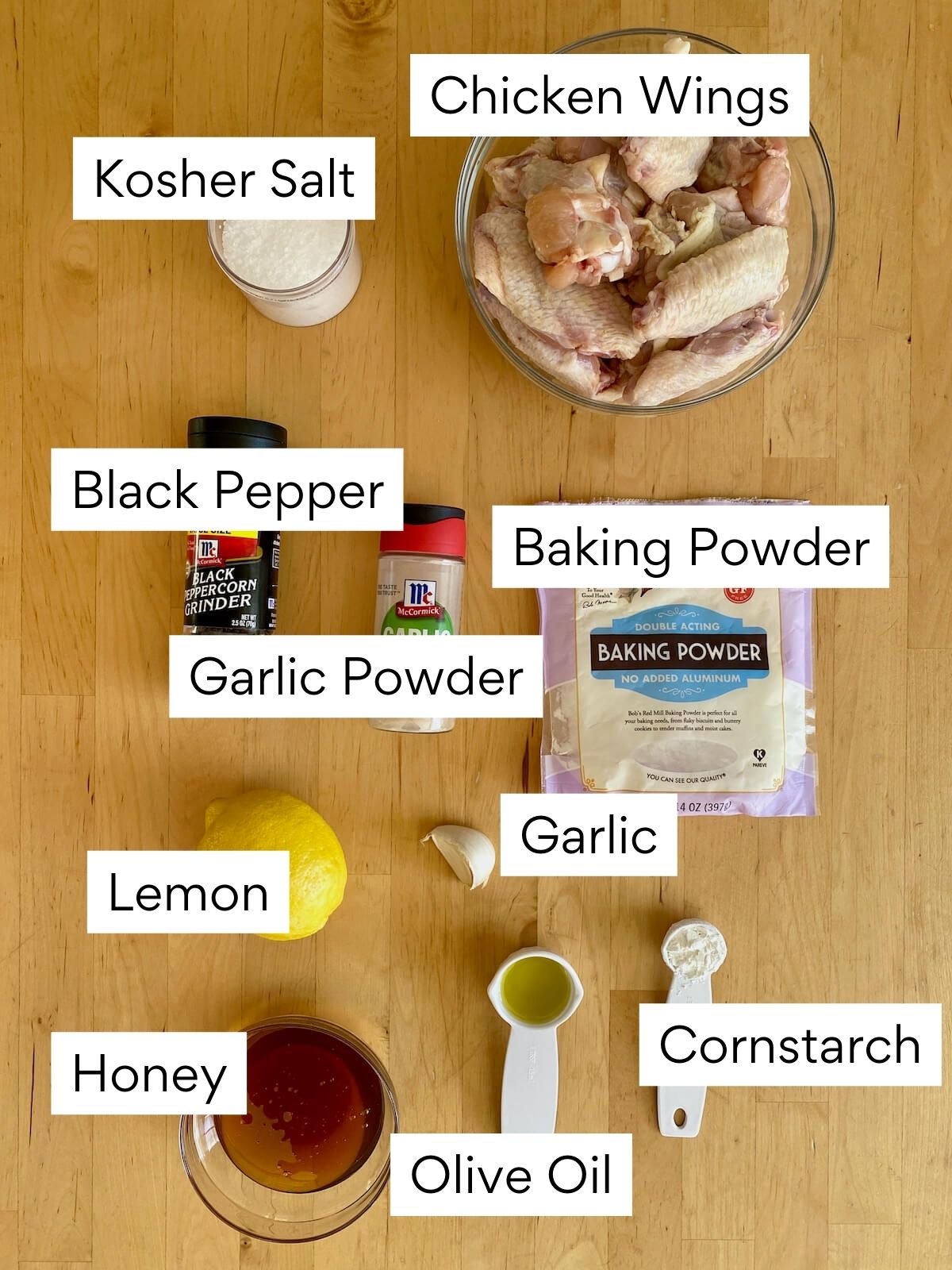The ingredients to make honey lemon pepper wings. Each ingredient is labeled with text. They include chicken wings, kosher salt, black pepper, baking powder, garlic powder, garlic, lemon, honey, olive oil, and cornstarch.