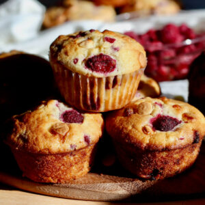 Three raspberry white chocolate muffins stacked in a triangle. Out of focus in the background are more muffins and a small bowl of raspberries.