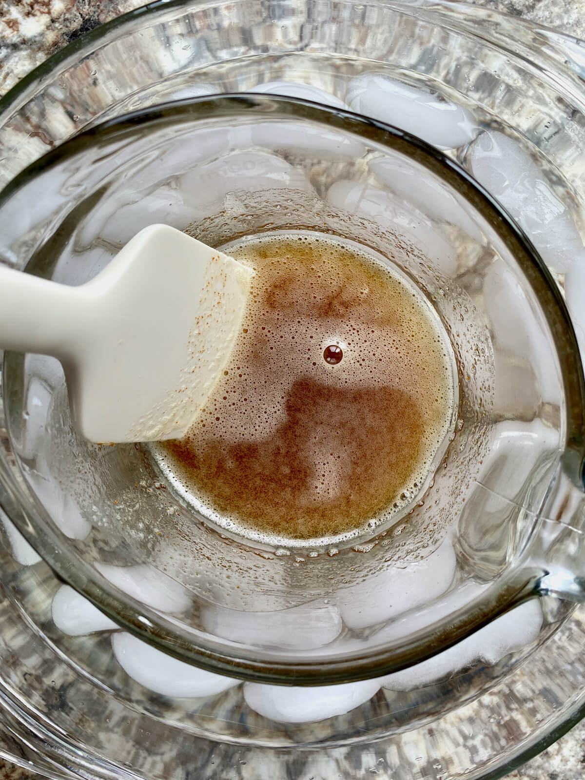 Brown butter in a glass measuring cup sitting inside a large bowl of ice water. There is a rubber spatula in the brown butter.
