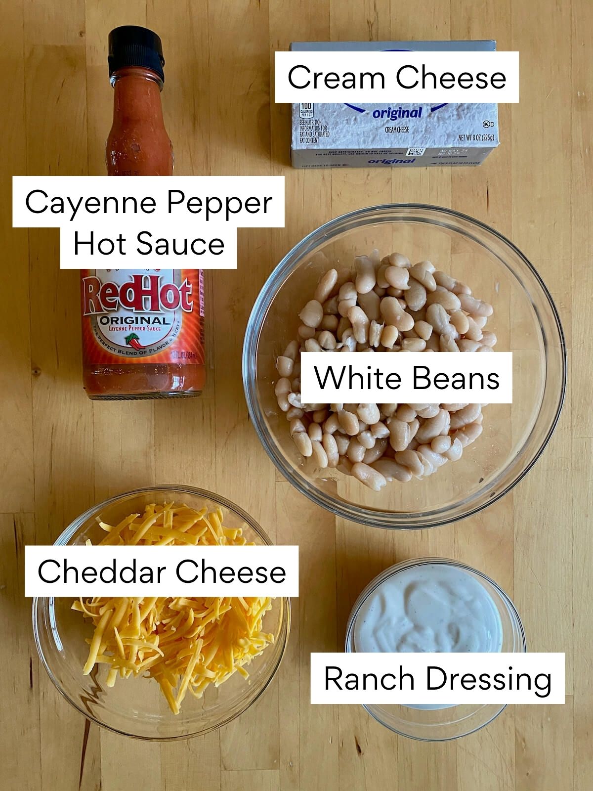 The ingredients to make white bean buffalo dip. Each ingredient is labeled with text. They include cream cheese, cayenne pepper hot sauce, white beans, cheddar cheese, and ranch dressing.
