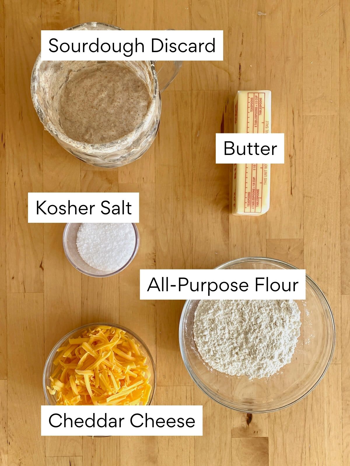 The ingredients to make sourdough discard cheese crackers. Each ingredient is labeled with text. They include sourdough discard, butter, kosher salt, all-purpose flour, and cheddar cheese.