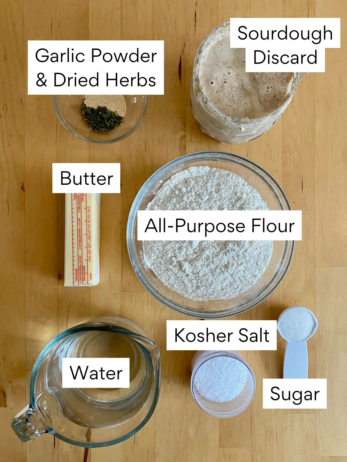 The ingredients to make sourdough garlic knots. Each ingredient is labeled with text. They include garlic powder and dried herbs, sourdough discard, butter, all-purpose flour, water, kosher salt, and sugar.