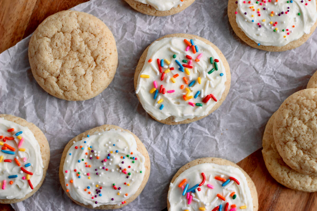 Several sourdough sugar cookies displayed on a piece of crinkled parchment paper on a wooden countertop. Some of the sugar cookies are decorated with frosting and rainbow sprinkles.